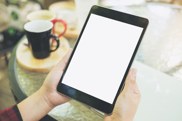 Mockup image of hands holding black tablet pc with blank white screen with coffee cups on glass table in modern cafe