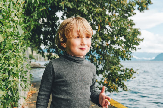 Outdoor portrait of funny little boy walking next to beautiful lake Geneva, wearing grey turtleneck shirt