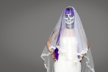 Portrait of woman looks at the camera with terrifying halloween skeleton makeup and purple wig bridal veil, wedding dress over gray background. Black wedding