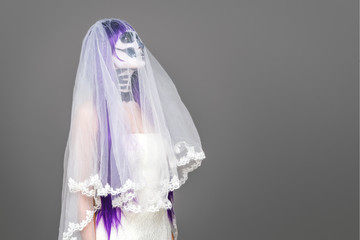 Portrait of woman looks upwards with terrifying halloween skeleton makeup and purple wig bridal veil, wedding dress over gray background. Black wedding. Copy space