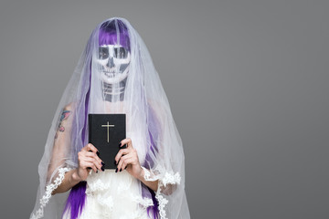 Portrait of woman looks at the camera with terrifying halloween skeleton makeup and purple wig bridal veil, wedding dress, holds the Holy Bible over gray background. Black wedding. Copy space
