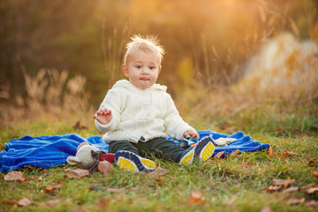 Little funny boy with sticking up hair sitting on a blue plaid on green lawn and playing with toys at sunset.
