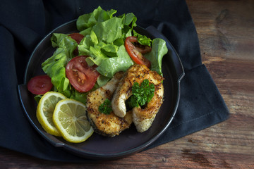 fried northern pike steaks with lemon, lettuce salad, tomatoes and parsley garnish on a black plate, healthy low carb meal on a dark wooden table, top view from above