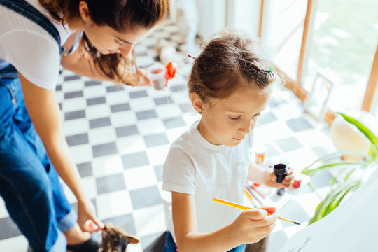 Happy family. Top view of little daughter dressed in white shirt and blue jeans painting on easel with a brush. Adult woman feeding the cat on background. New housing, art and family leisure concept.