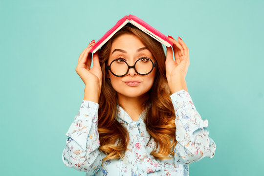 Beautiful young student holding book on her head and wearing glasses over blue background