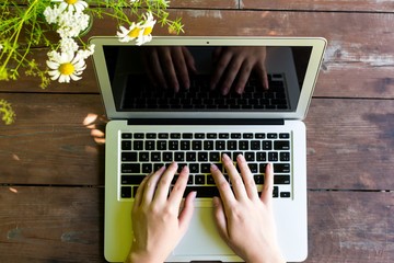 Close up view of female hands on keyboard of laptop. Woman works with laptop on wooden table at garden  and flowers near