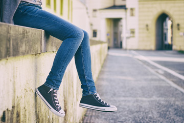 Young female tourist sitting on a wall in a historic european city. A woman wearing jeans and new sneakers.  - obrazy, fototapety, plakaty