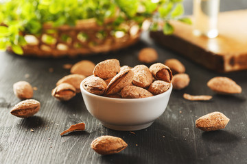 Almond in shell in a white ceramic bowl on a black wooden table with greenery behind it