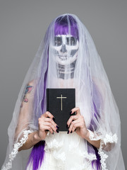 Portrait of woman looks at the camera with terrifying halloween skeleton makeup and purple wig bridal veil, wedding dress, holds the Holy Bible over gray background. Black wedding