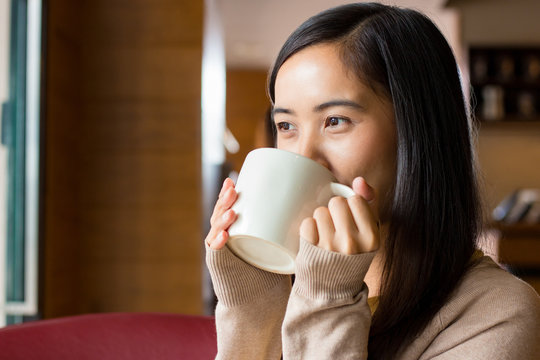 Asian woman drinking coffee from white coffee cup with blurry background of coffee shop
