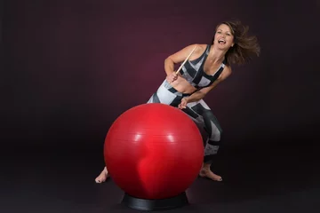 Reifere Frau im Sport Outfit trommelt mit Drum Sticks auf einem roten Gymnastikball vor einem dunklen Hintergrund © Frozen Action