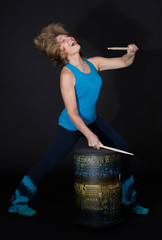 Reifere Frau im blauen Sport Outfit trommelt mit Drum Sticks auf einer Trommel vor einem dunklen Hintergrund