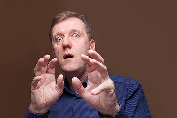 A very frightened man with wide open eyes and an open mouth is protected by raised hands