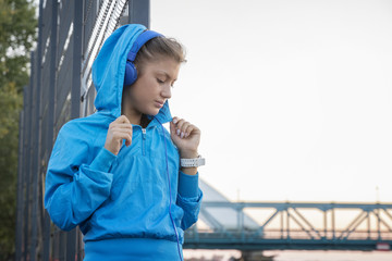 Young fitness girl standing with headphones on her head and bridge on background