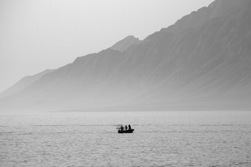 Picturing the seascape in the morning with the background is Rocky mountain. A silhouette of fishermen on the boat while fishing. The picture is in black and white.