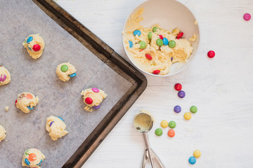 Cookie dough with colorful chocolate candies in bowl next to dough scooper and scoops of cookie dough on baking sheet