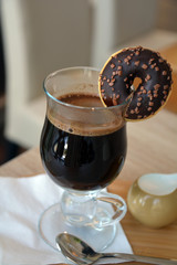 Black coffee on a glass cup and small donut on a wooden table.
