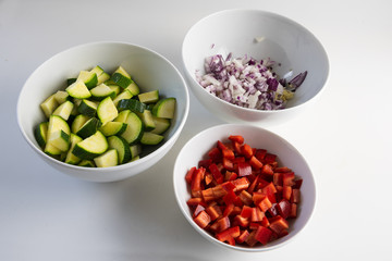 sliced vegetables, zucchini, peppers and onions in white bowls on a light gray background