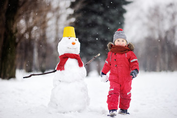 Fototapeta na wymiar Little boy in red winter clothes having fun with snowman in winter park