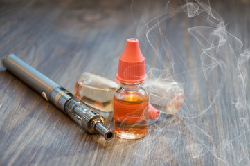 An electronic cigarette, a vape, and several bottles of liquid for refilling lie on a wooden background. with smoke around