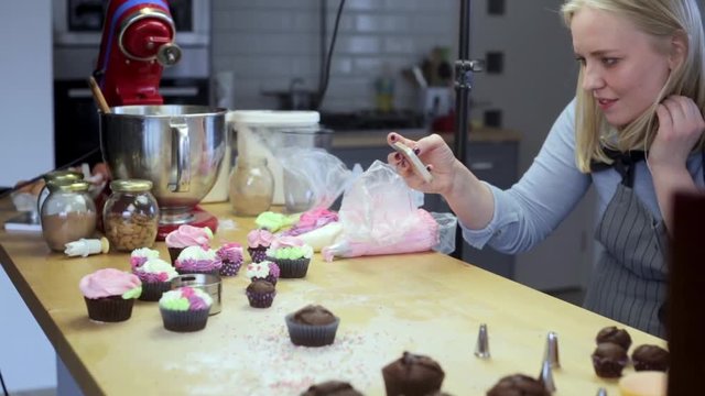 Young woman taking photos of cupcakes she has bakes in the kitchen. Female using smartphone to take pictures of desserts