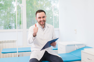 Cheerful young doctor holding a clipboard and gesturing with his hand at hospital ward