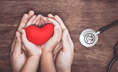 Red heart on woman and child hands and with doctor's stethoscope on wooden background. Healthcare...
