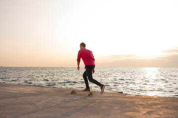 skater in red shirt and blue jeans riding on concrete pier on longboard during sunrise, sea or ocean background  