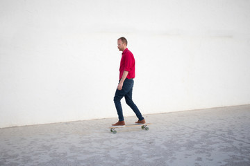 Fototapeta na wymiar Young bearded man riding on skateboard, hipster with longboard in red shirt and blue jeans urban background 