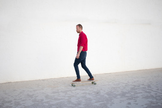 Young bearded man riding on skateboard, hipster with longboard in red 
shirt and blue jeans urban background  