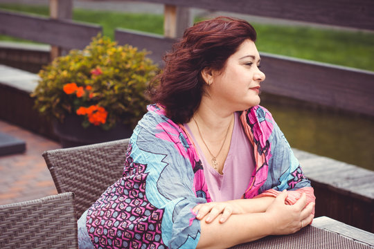 Mature woman in bright scarf, size plus american or European appearance sitting in a restaurant drinks coffee at autumn day. Lady with excess weight, stylishly dressed in cafe waiting for a friend