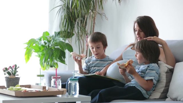 Happy young family, pregnant mother and two boys, eating tasty pizza at home, sitting on the sofa, reading a book and having a laugh
