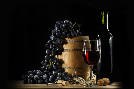 A still life includes red wine in glass, wine bottle, wooden barrel, grape