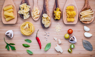Italian food concept .Various kind of pasta in wooden spoons with ingredients sweet basil ,tomato ,garlic ,parsley ,bay leaves ,pepper ,champignon and parmesan cheese on bamboo cutting board flat lay.