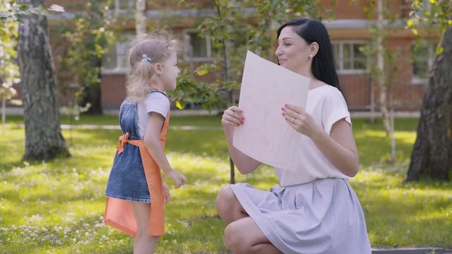 Small joyful girl with two tails and in an orange apron runs to mother, holding a painted picture. Baby gives mother a picture drawn in the kindergarten, mom kisses daughter. A gift to parents.