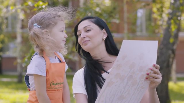 Small joyful girl with two tails and in an orange apron runs to mother, holding a painted picture. Baby gives mother a picture drawn in the kindergarten, mom kisses daughter. A gift to parents.