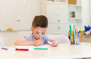 child preschooler boy drawing on booklet ,while sitting on the desk in the classroom