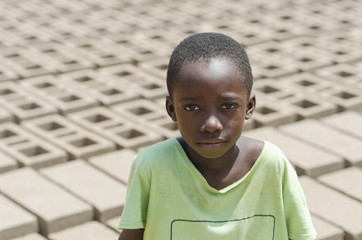 Black child sitting outdoors as a child labor concept - Isolated in front of a lot of bricks to build