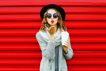 Fashion portrait pretty woman with coffee cup blows red lips sends an air kiss on a red background