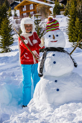 Funny happy girl in a red knitted nordic sweater and warm hat  playing with a snowman. Kids play outdoors in winter holidays, children having fun at Christmas time. Child building snowman.