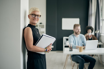 Businesswoman holding documents and standing against the wall in modern office