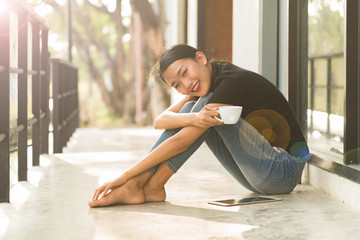 Fototapeta na wymiar Asian woman smiling happy drinking coffee in sun sitting outdoor home in sunshine light enjoying her morning coffee.