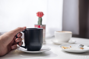 Closeup image of a hand holding a black cup of hot coffee and white cup , plate and cactus on vintage wooden table in minimalism cafe