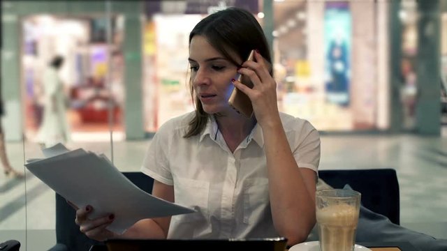 Young businesswoman with documents talking on cellphone in cafe by window
