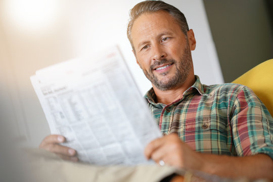 Cheerful mature man reading newspaper at home