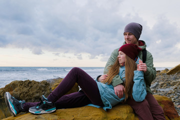 young couple sitting on rocks on the shore, hugging and looking at sea