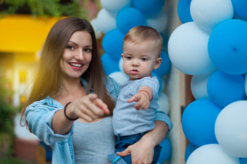 Portrait of young beautiful mother with her year-old baby boy.Both point the finger in front of him on the camera on balloons background
