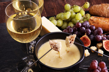 Gourmet Swiss fondue dinner on a winter evening with assorted cheeses on a board alongside a heated pot of cheese fondue with two forks dipping bread and white wine behind in a tavern or restaurant