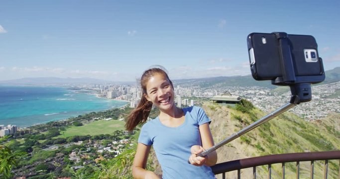 Cheerful hiker taking selfie using smart phone using selfie stick by Waikiki Beach and Honolulu. Happy woman is on vacation at Diamond Head State Monument. She is photographing at Oahu, Hawaii, USA.