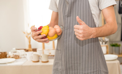 young attractive man at home kitchen happy leaning on table , vegetables and knife wearing Gray apron giving thumb up in learning cooking  , lifestyle at home concept,selective focus on hand thumb up